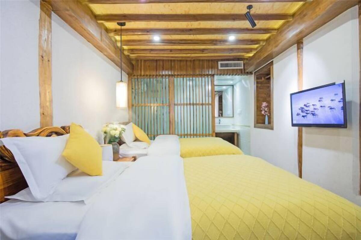 A hotel room with wooden beams, a bed with white and yellow bedding, a television on the wall, and a window with a view.