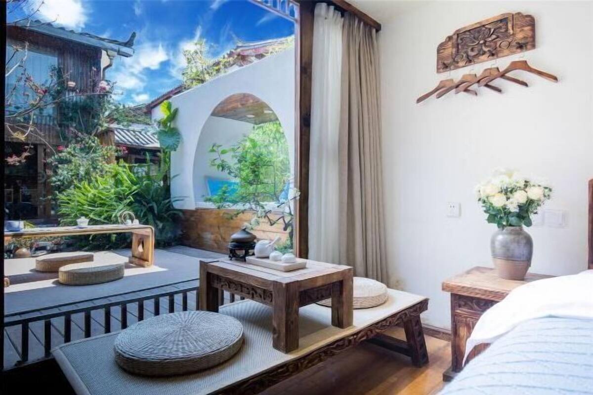 A room with a traditional wooden table, a small bench, and a bed. There is a view of a garden through an archway.