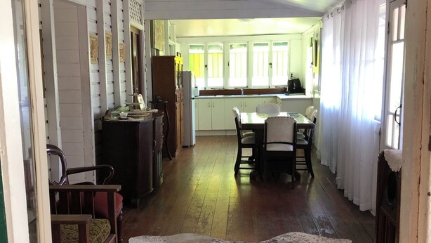 A spacious room with a wooden floor, a dining table with chairs, a large white rug, and a kitchen area with cabinets and a window.