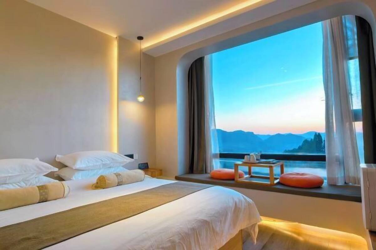 Quarto de hotel com uma cama, uma mesa pequena e vista para montanhas pela janela.
