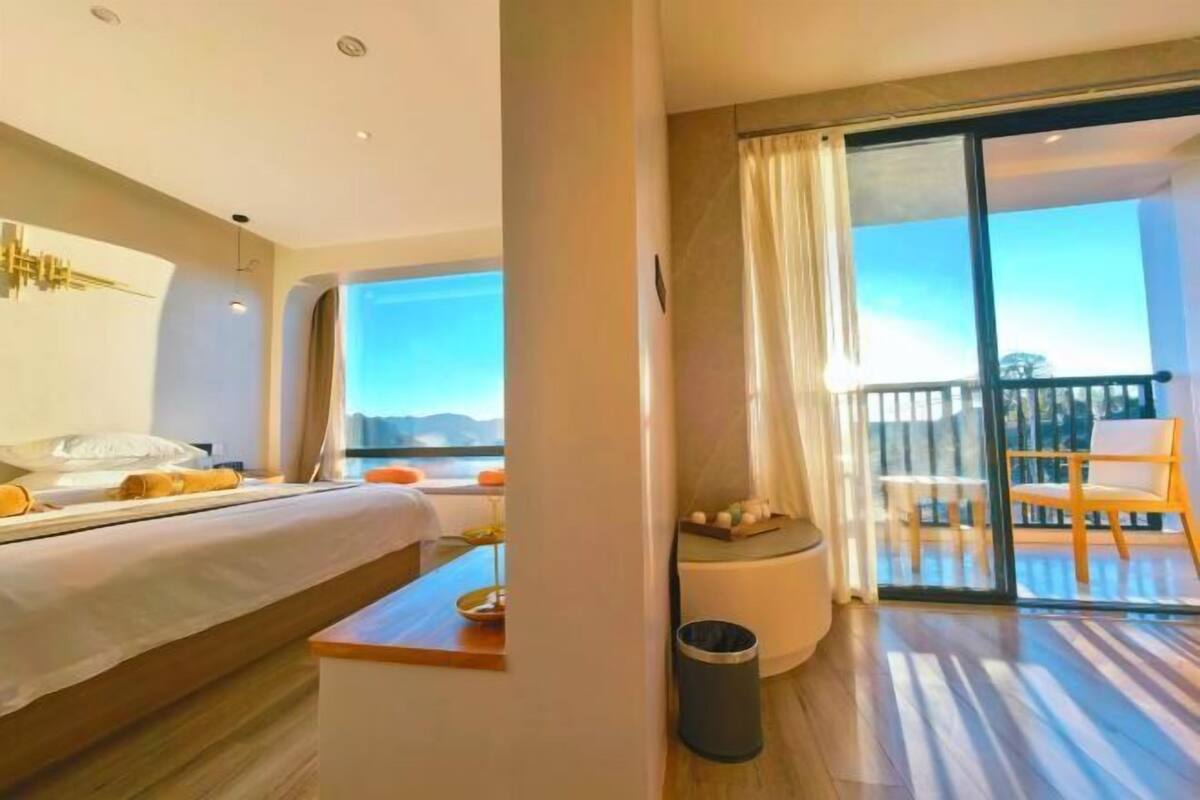 Quarto de hotel com cama, uma mesinha, um cesto de lixo e varanda com cadeiras.