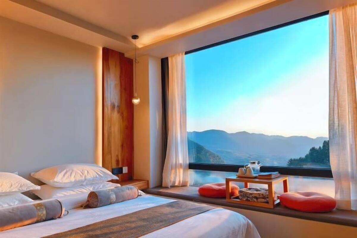 Quarto com uma grande janela, cama com travesseiros, mesa de cabeceira com abajur e vista para montanhas.