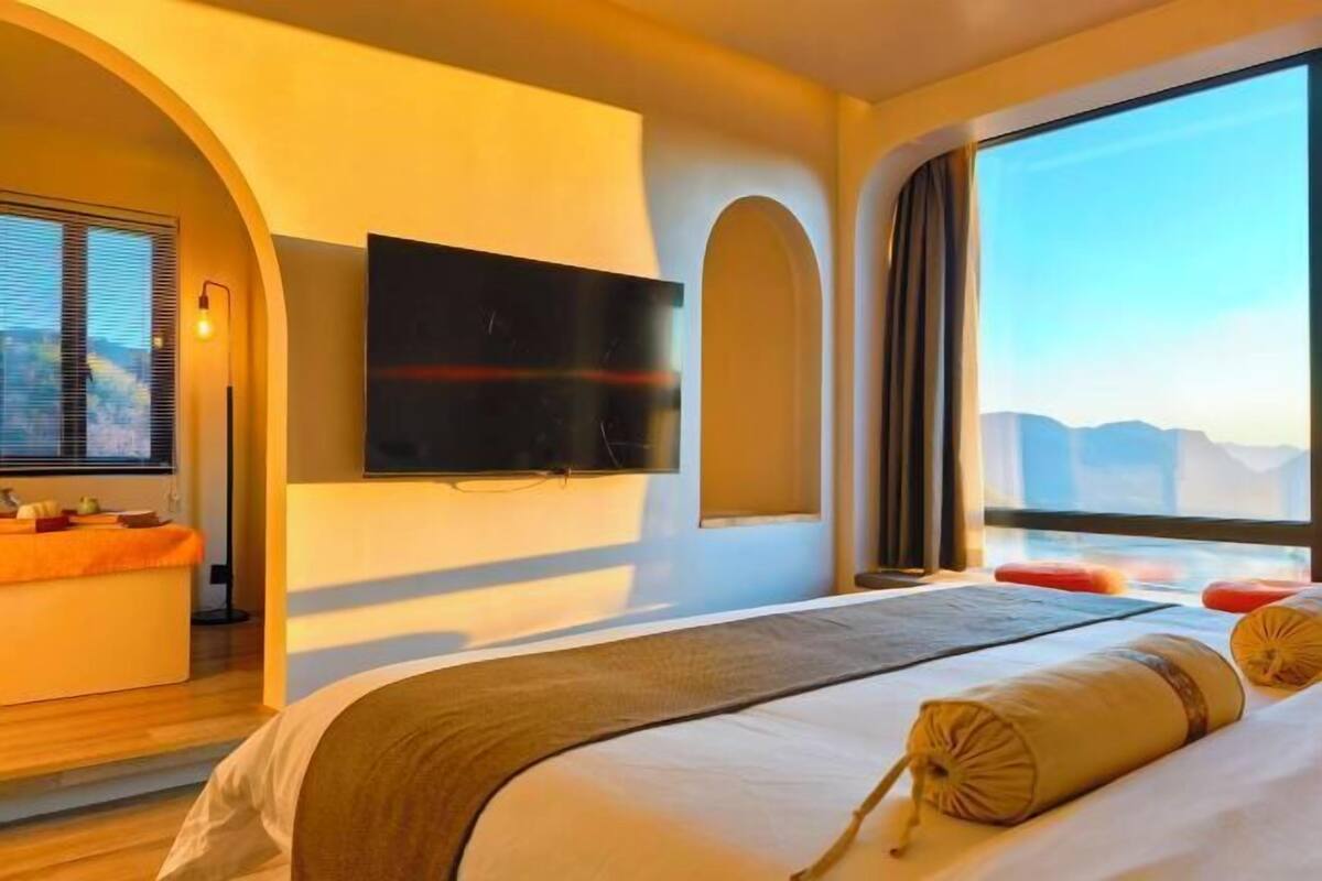 Quarto de hotel com cama, televisão na parede, janela com vista e um pequeno espaço de estar com mesa e cadeiras.