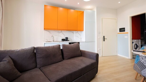 Uma sala de estar moderna com sofá, cozinha com armários laranja, máquina de lavar e porta que dá para outro cômodo.