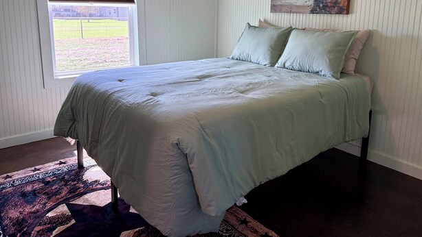 A bedroom with a bed, a window with blinds, a painting on the wall, and a patterned rug.