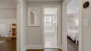 Un couloir menant à une salle de bain équipée d’une baignoire, dotée d’un plancher en bois et d’une porte blanche.