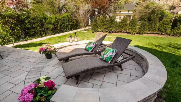 Une terrasse circulaire en pierre, agrémentée de deux fauteuils de jardin en osier et d’une petite table, le tout entouré d’une végétation luxuriante et d’une pelouse bien entretenue.
