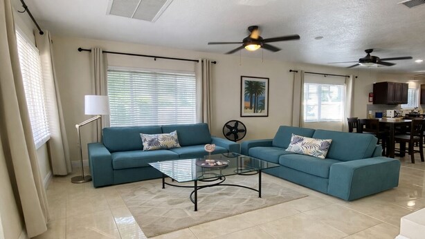 A living room with two sofas, a coffee table, ceiling fans, and a kitchen area in the background. - Entire Private 2BR 2 Bath Townhouse Near Seven Mile Beach w/ Balcony + Garden (George Town)