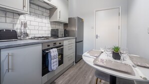 A modern kitchen with white cabinets, a stainless steel refrigerator, a microwave, and a stove. There is a dining table with a plant and wine glasses. - Modern Flat in Waterloo - Central London (London)
