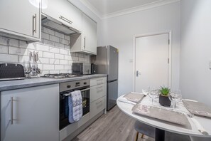 A modern kitchen with white cabinets, a stainless steel refrigerator, a microwave, and a stove. There is a dining table with a plant and wine glasses. - Modern Flat in Waterloo - Central London (London)