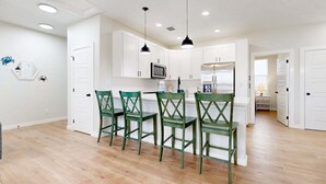 A kitchen with white cabinets, a microwave, and a refrigerator. There are four green chairs around a small island. The floor is wooden, and the ceiling has recessed lighting. - MG303l, Shared Heated Pool, Pet Friendly (Port Aransas)