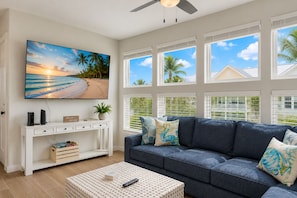 Ein Wohnzimmer mit großem Fernseher, einem weißen Konsolentisch, einem blauen Sofa und Blick auf den Strand durch große Fenster.