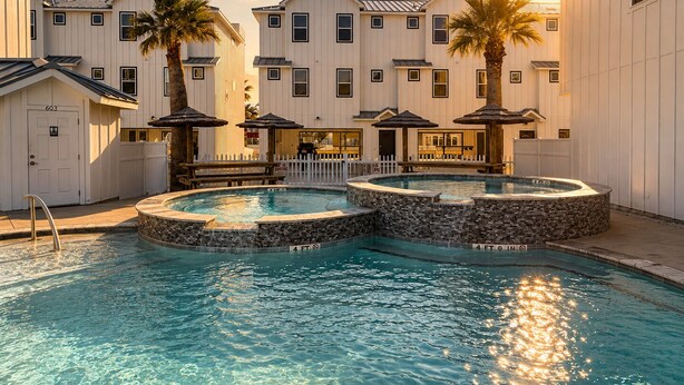 A pool area with two hot tubs, a dining area, and a building with a white facade. - MG303l, Shared Heated Pool, Pet Friendly (Port Aransas)