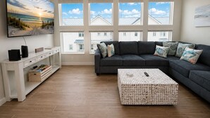 A living room with a sectional sofa, a coffee table, a TV, and a large window with a view of buildings. - MG303l, Shared Heated Pool, Pet Friendly (Port Aransas)