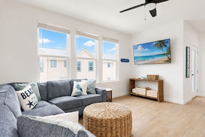 A living room with a grey sofa, wicker ottoman, and a beach-themed painting. - MG304, Shared Heated Pool, Golf Cary Included, Pet Friendly (Port Aransas)