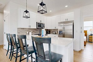 A modern kitchen with white cabinets, a central island, bar stools, a microwave, a refrigerator, and pendant lighting. - MG304, Shared Heated Pool, Golf Cary Included, Pet Friendly (Port Aransas)