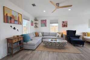 Un salon moderne avec un canapé d’angle gris, une table basse ronde, un fauteuil foncé et un ventilateur de plafond.