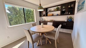 A dining area with a round table, four chairs, a hanging light fixture, dark wooden cabinets, and a window with a view of trees.
