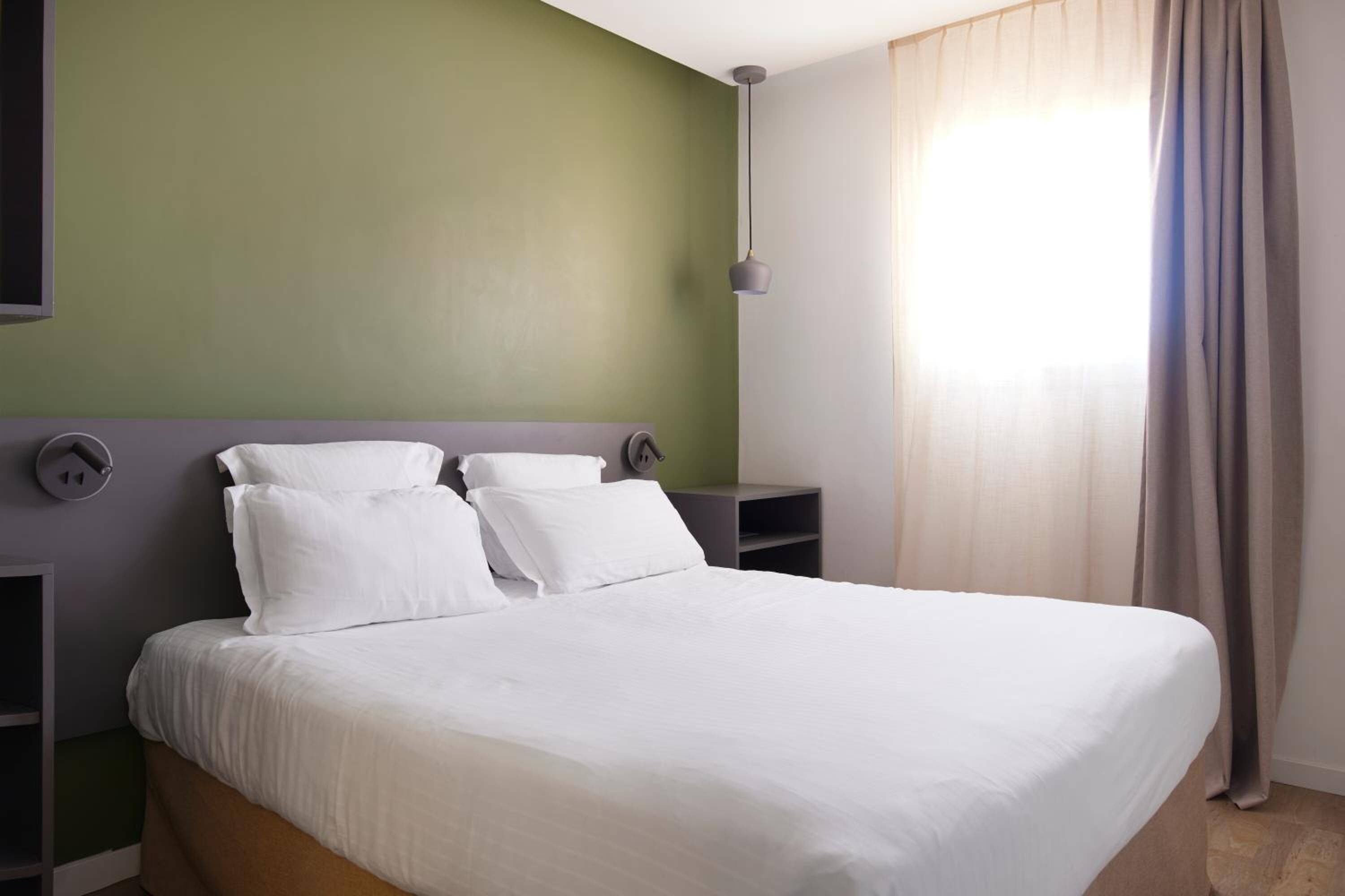 A hotel room with a bed, bedside tables, a wall-mounted lamp, and a window with curtains.