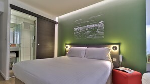 Een moderne slaapkamer met een groot bed, een rode poef, een nachtkastje met een lamp en een groene accentmuur.