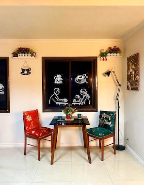 A dining area with two wooden chairs, a small table, a potted plant, and a window with a coffee-themed artwork.