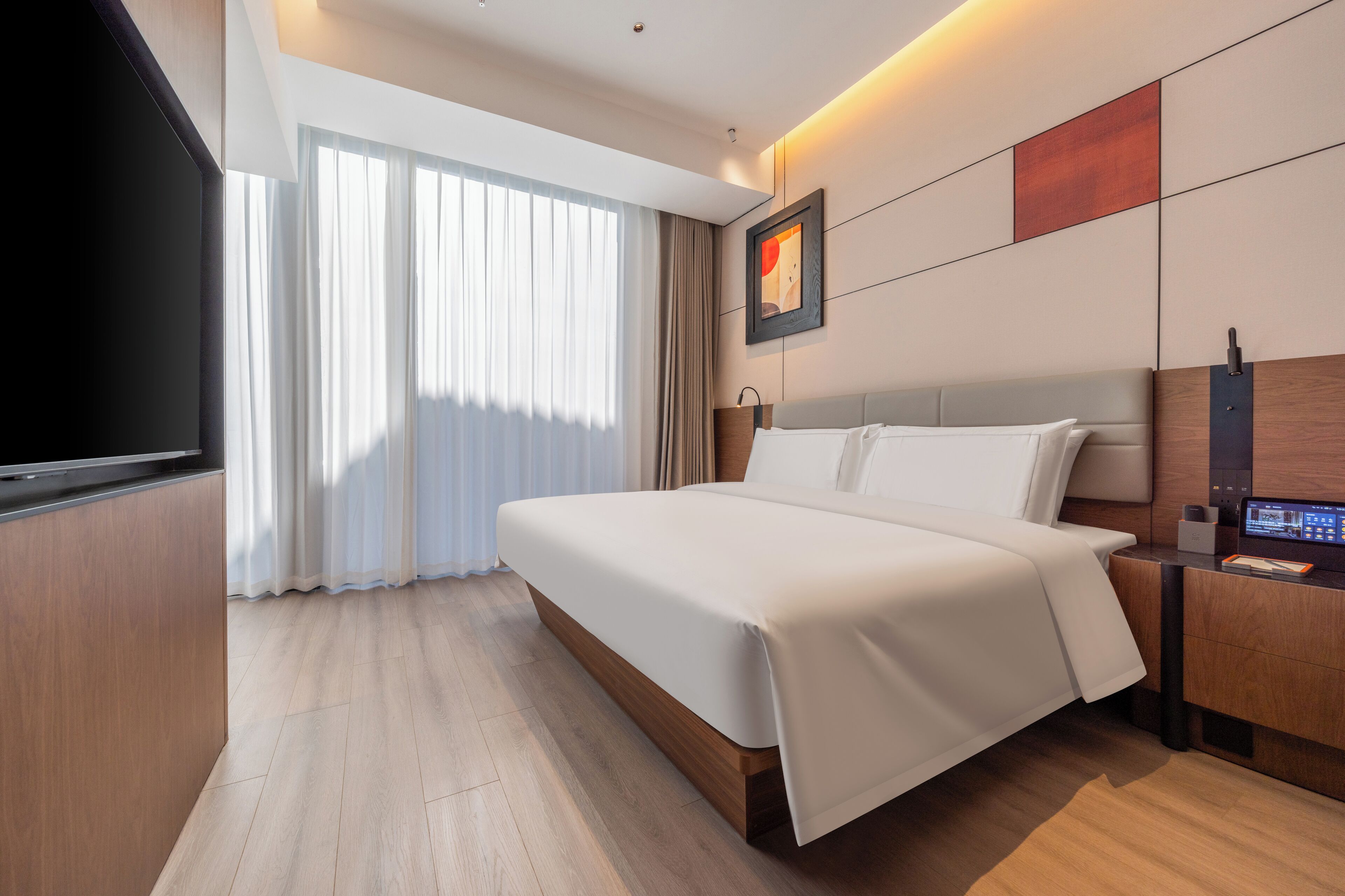 A modern hotel room with a large bed, a flat-screen TV, a desk with a computer, and a window with curtains.