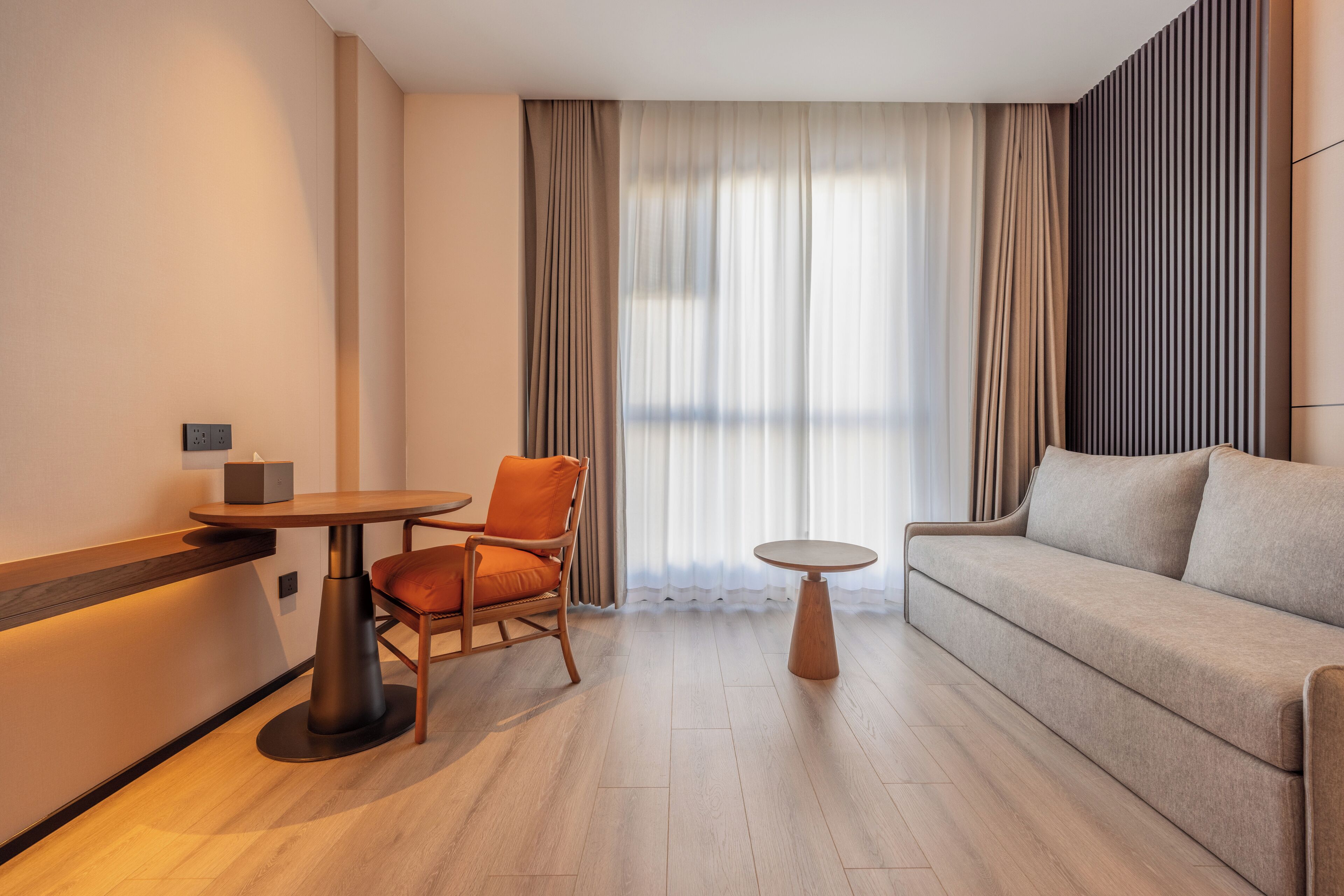 Una habitación de hotel moderna con piso de madera, un sofá, una silla, una pequeña mesa redonda y un gran ventanal con cortinas.