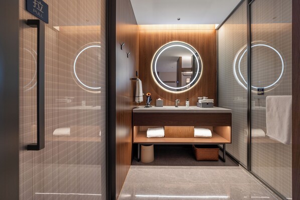 A modern bathroom with a wooden vanity, a round mirror with illuminated edges, and a glass shower enclosure.