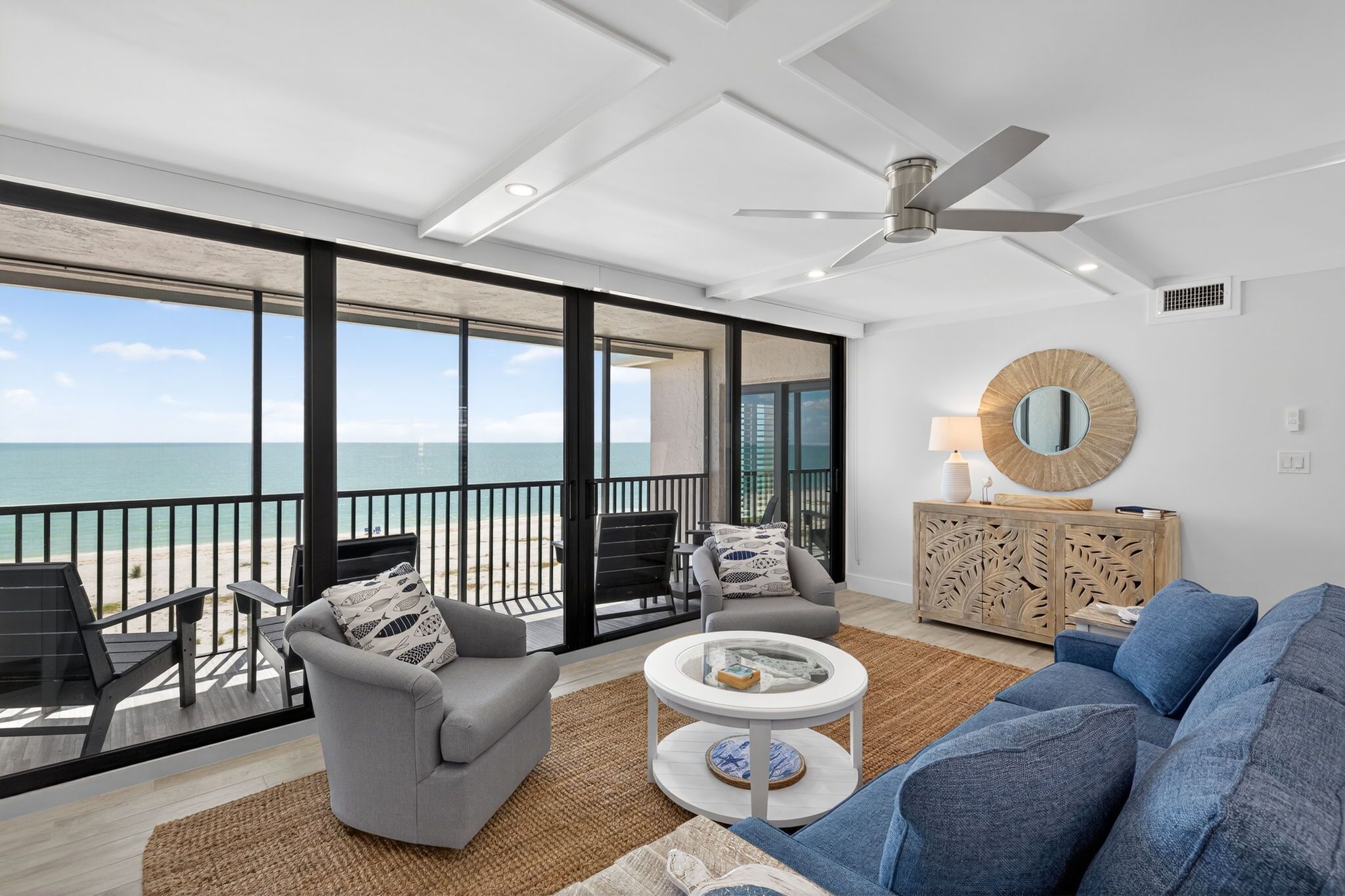 A spacious living area with a balcony overlooking the ocean, a ceiling fan, and a decorative wooden cabinet.