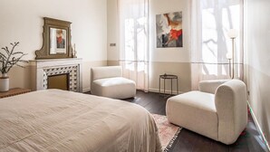 Une chambre d’hôtel avec un lit, deux fauteuils, une cheminée et des œuvres d’art aux murs.