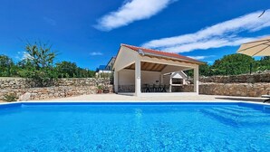 Ein Schwimmbecken mit überdachter Terrasse, Steinmauern und blauem Himmel.
