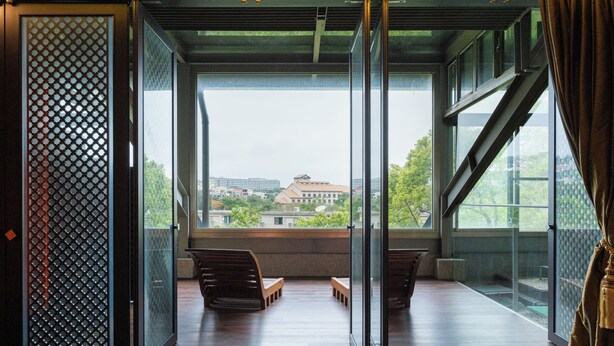 Un ambiente interno con vista su edifici e alberi attraverso ampie finestre, un’area salotto con panche e una porta scorrevole in vetro.