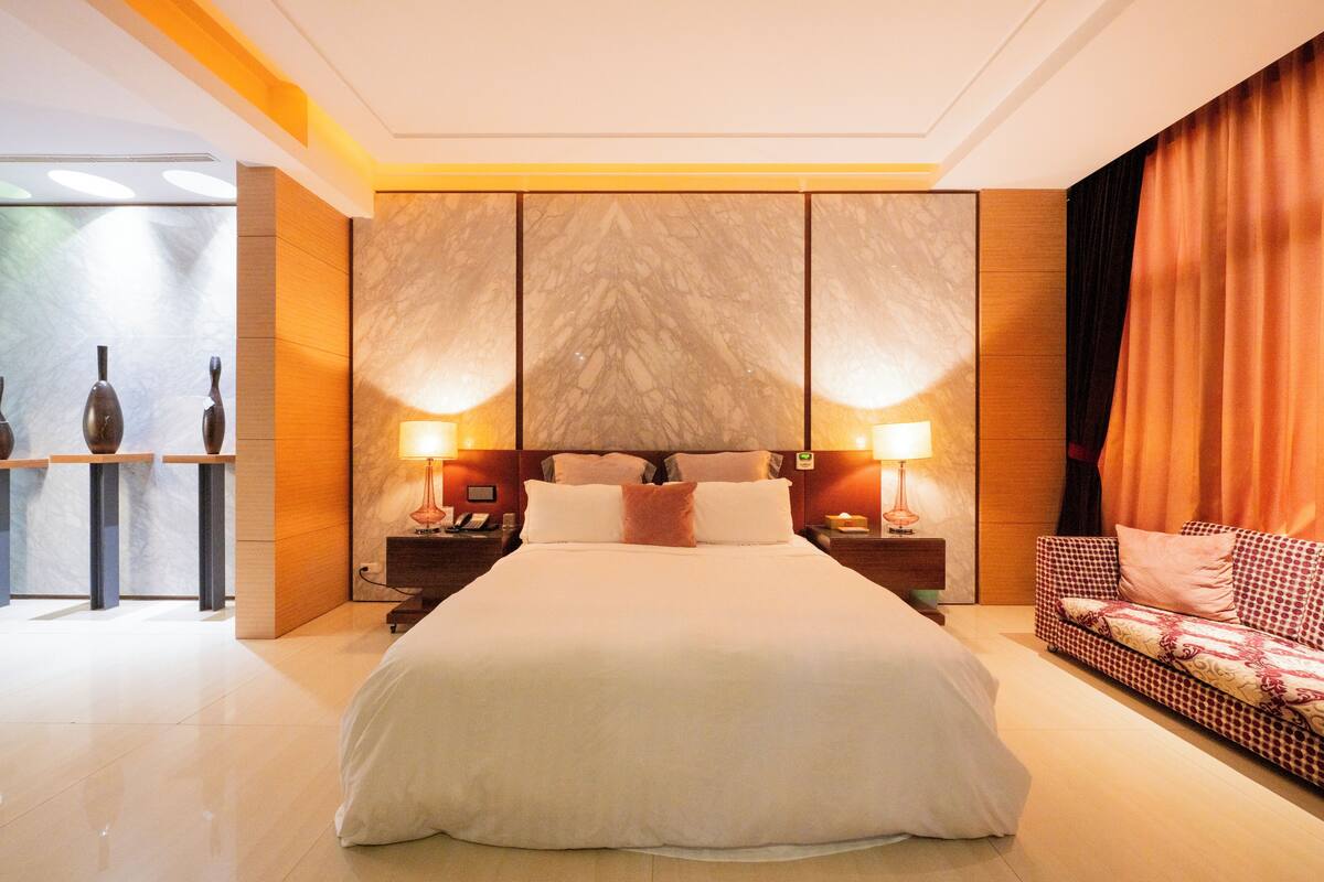 A hotel room with a large bed, two bedside tables with lamps, a sofa, and a decorative wall panel.