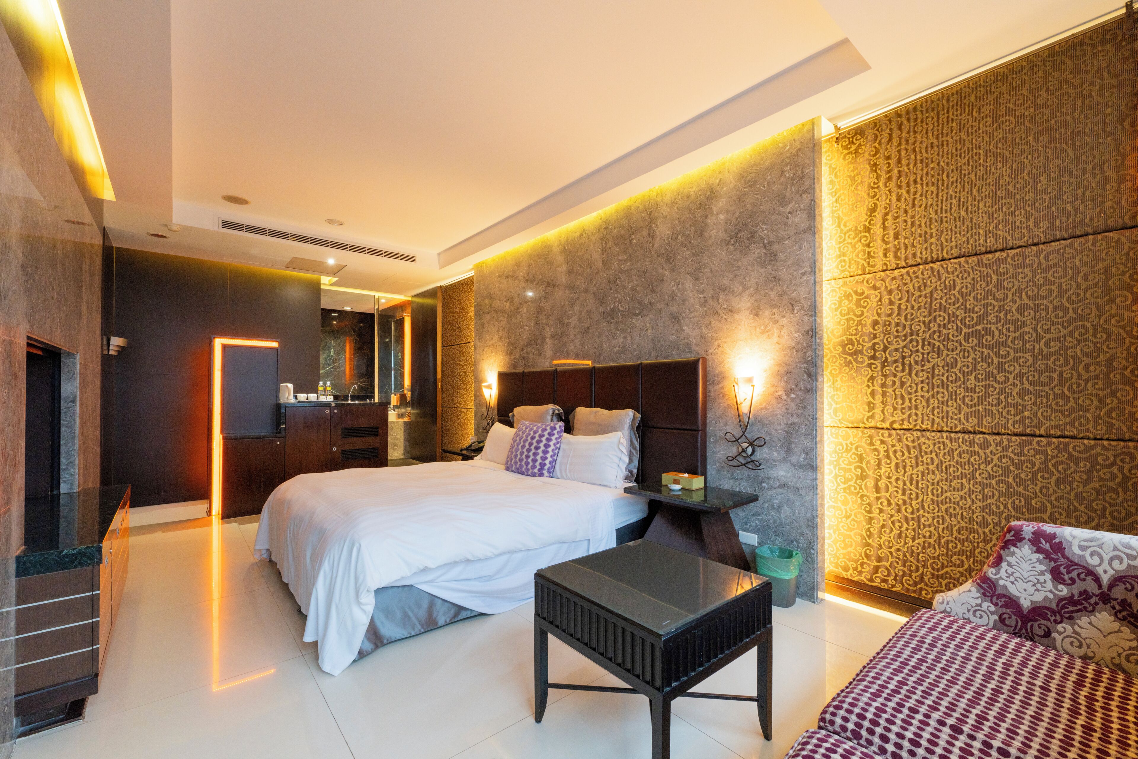 Une chambre d’hôtel moderne avec un grand lit, une petite table, un canapé, une salle de bain avec un miroir et un mur décoré de motifs.