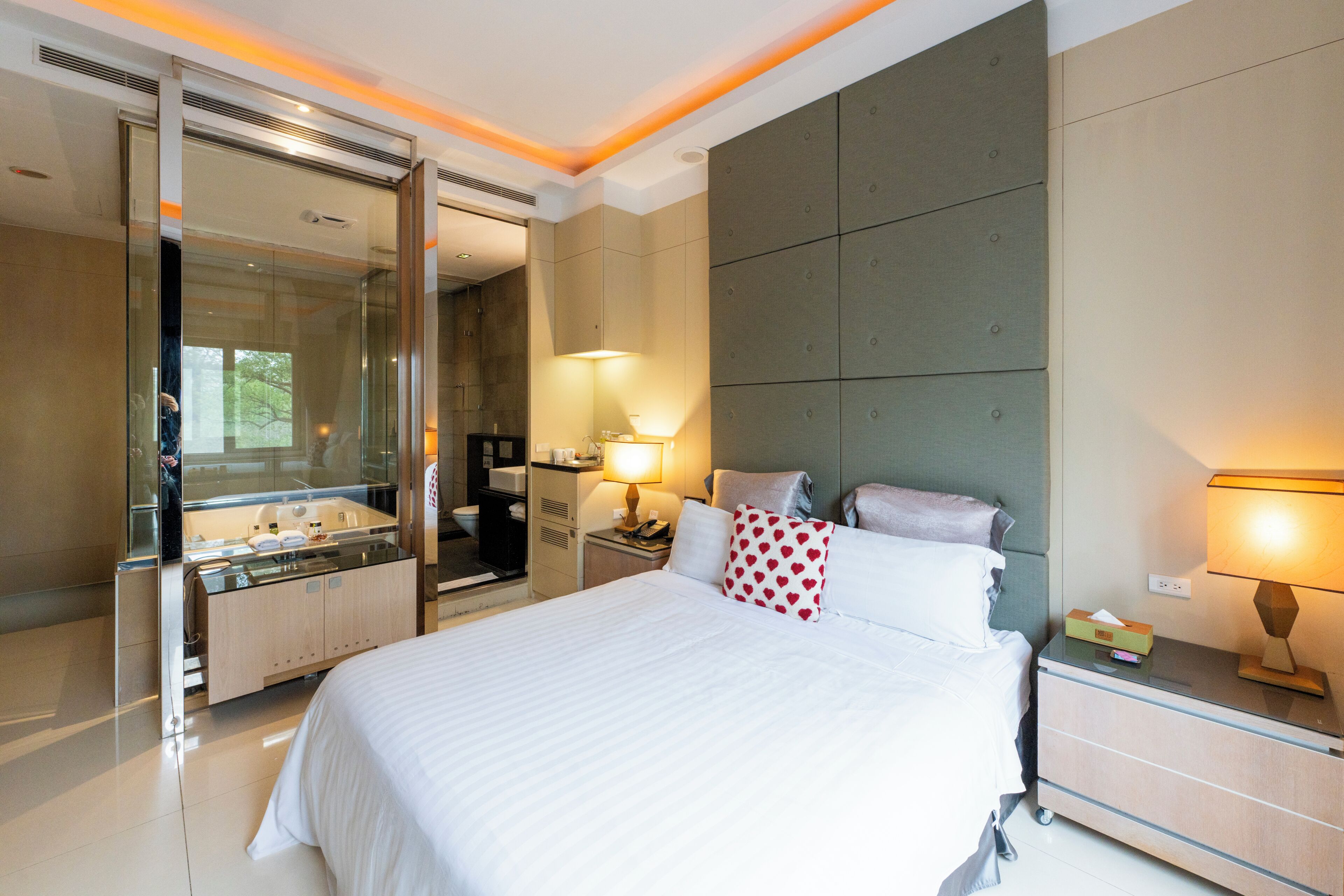 A modern hotel room with a large bed, a bedside table with a lamp, a nightstand, a mirror, and a bathroom with a glass shower enclosure.