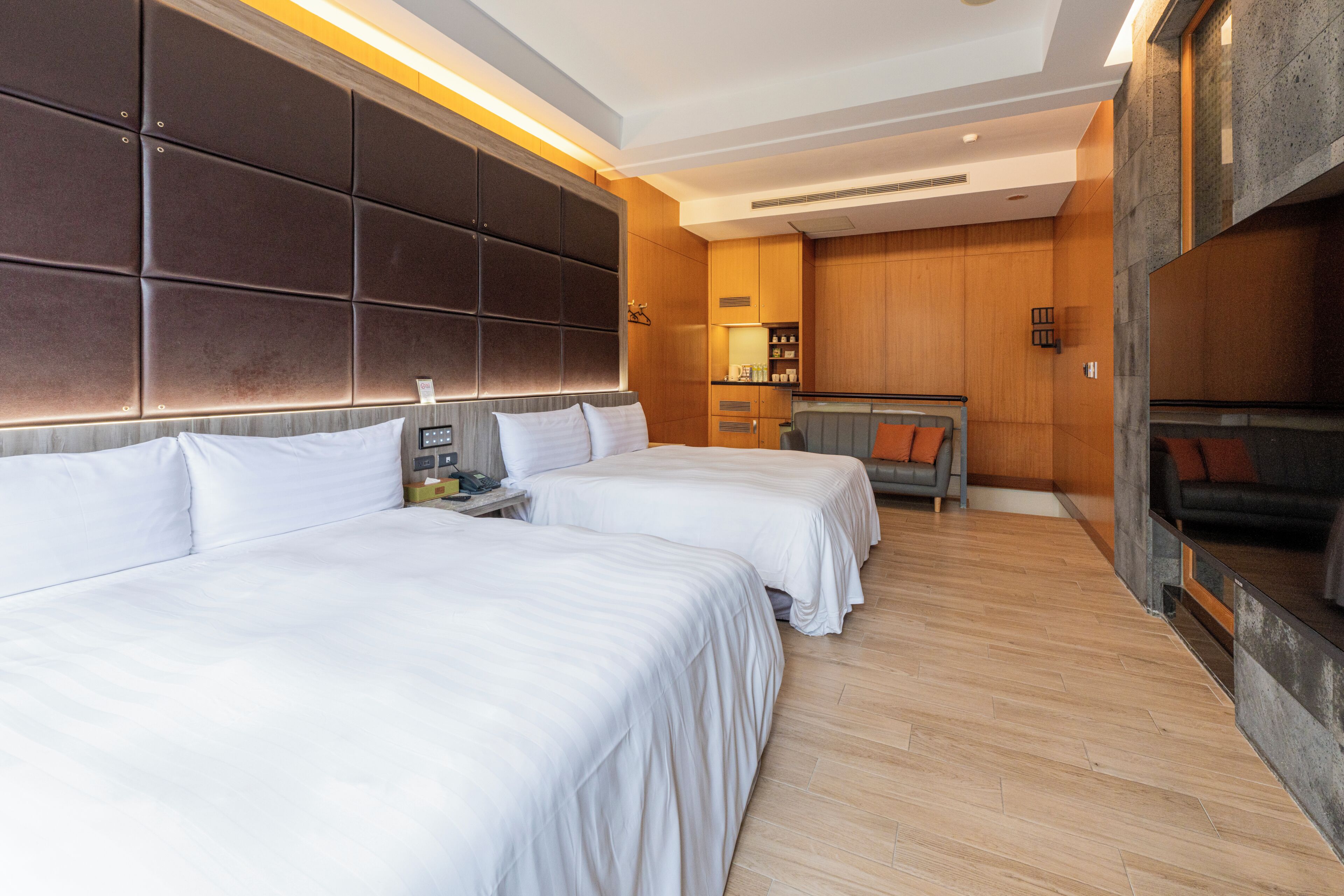 Habitación de hotel con dos camas, un sofá, un televisor, un escritorio con lámpara y un panel de pared de madera.