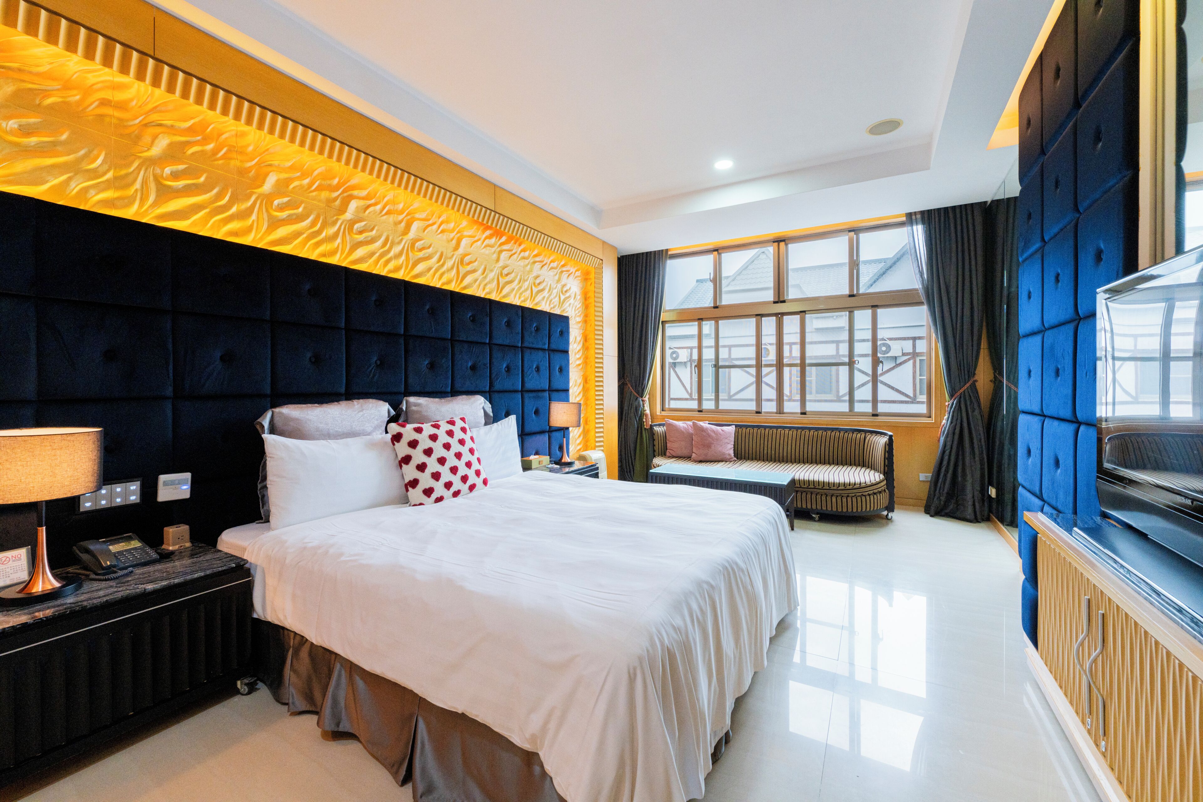 A hotel room with a large bed, a bedside table with a lamp, a window with curtains, and a seating area with a sofa and armchair.