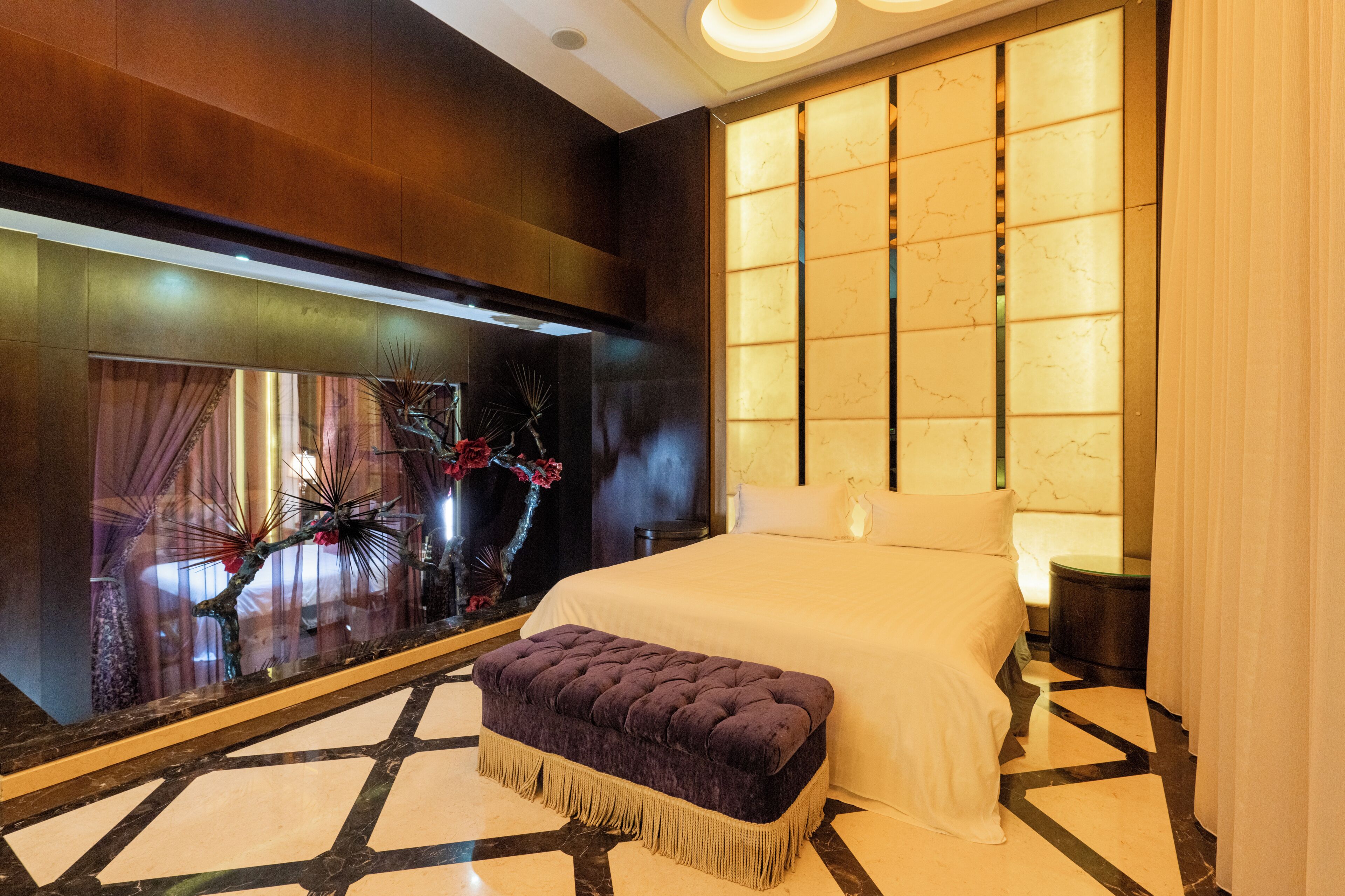 A hotel room with a large bed, a decorative wall with a reflective surface, and a small bench.