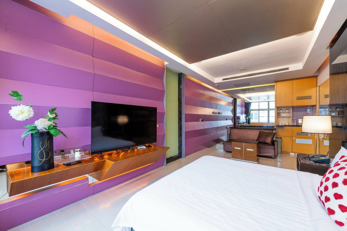 A modern hotel room with a large bed, a flat-screen TV, a wooden console table, a vase with flowers, and a kitchenette area.