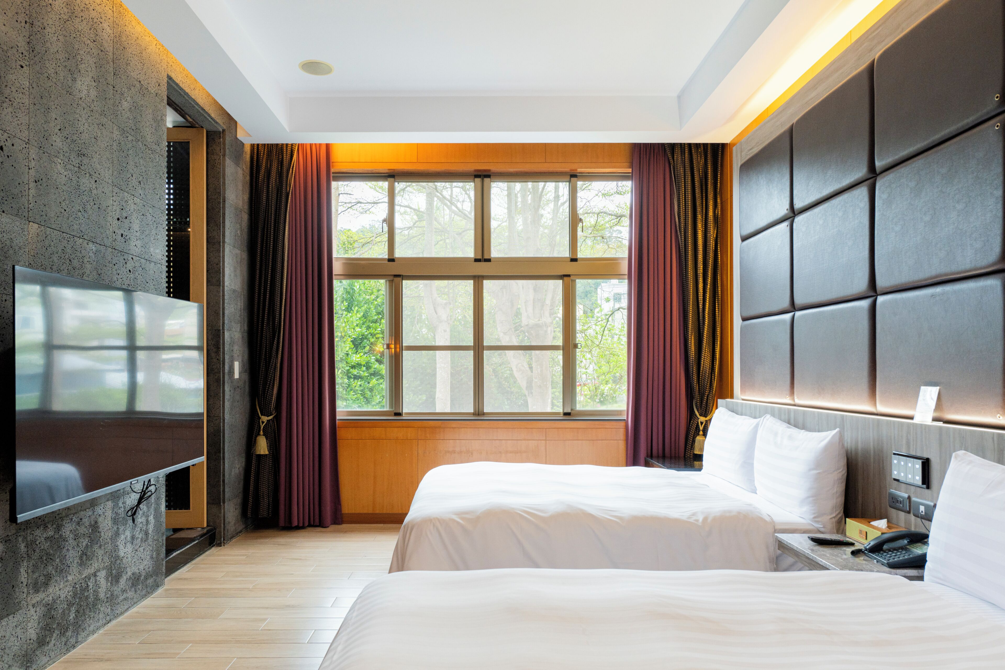 A hotel room with a large window, a flat-screen TV, a bed, and a bedside table.