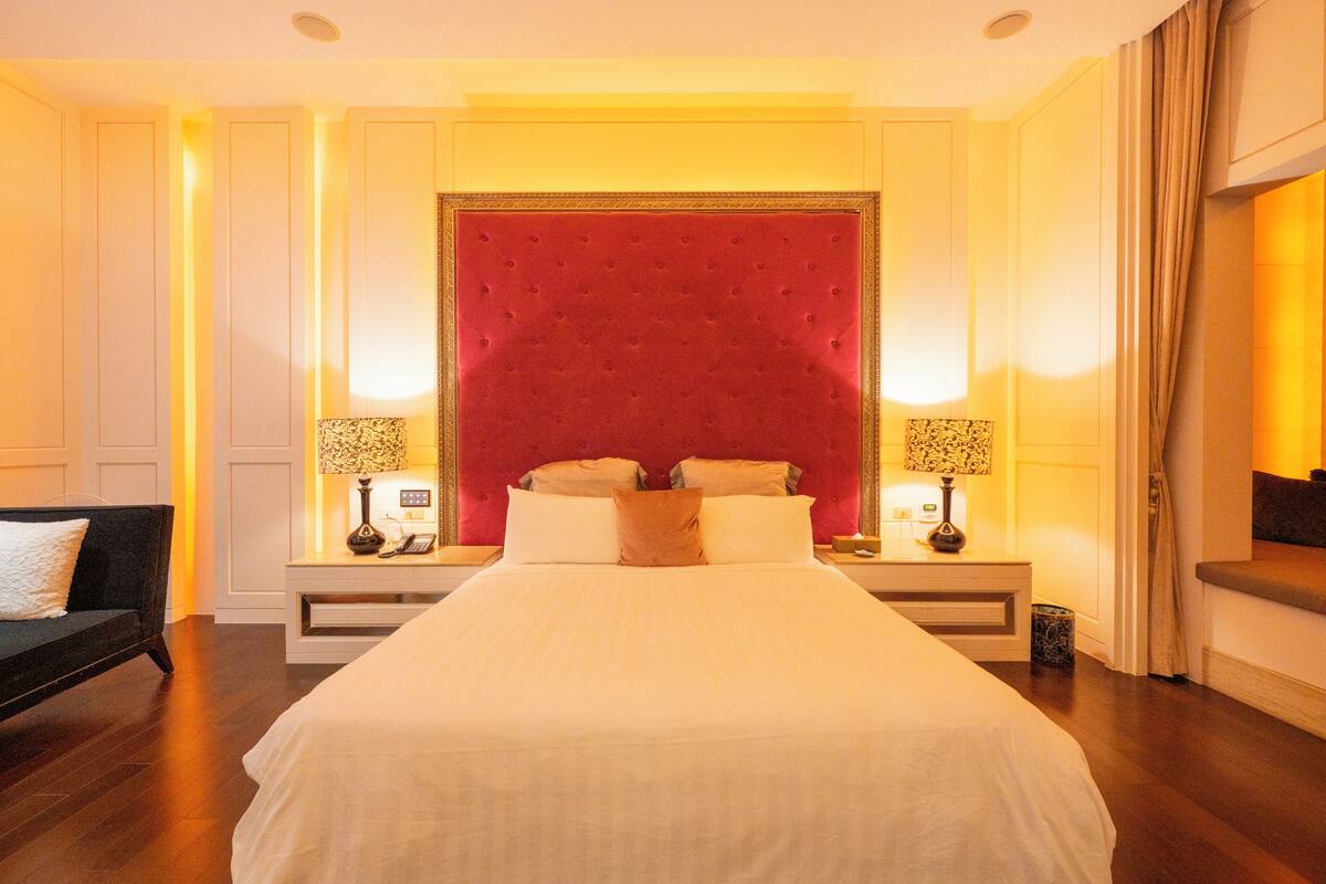 A hotel room with a large bed, two bedside tables, a nightstand, a sofa, and a red upholstered headboard.