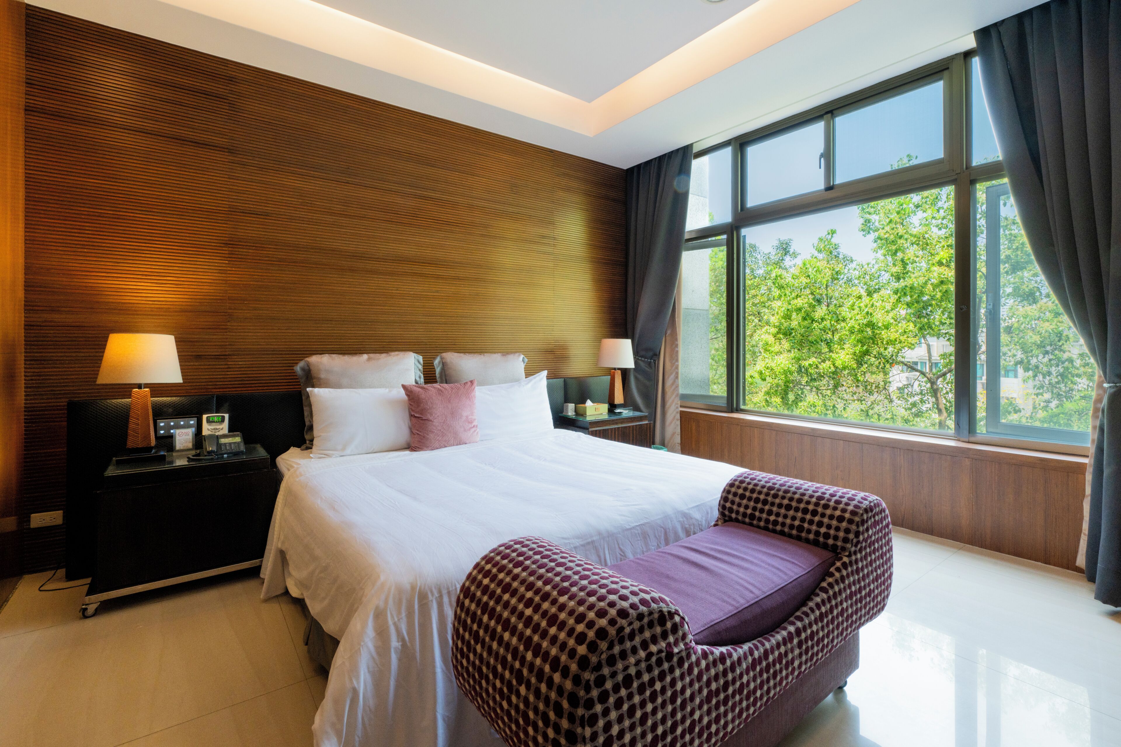 A modern hotel room with a large bed, bedside tables, a nightstand, a chair, and a large window offering a view of trees.