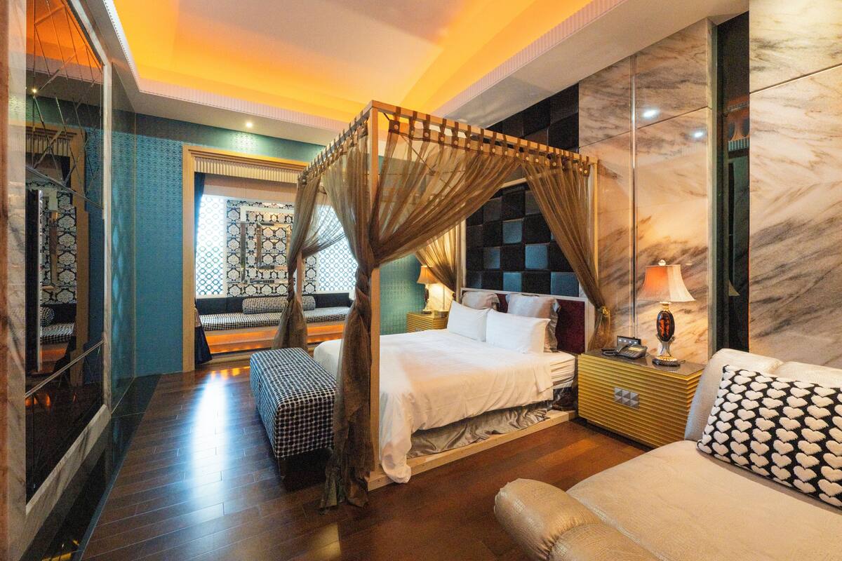 A bedroom with a canopy bed, a sofa, a bedside table with a lamp, and a patterned wall.
