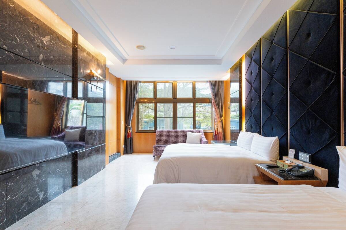 A hotel room with a large bed, a smaller bed, a sofa, a desk with a phone, and a view of the outdoors through large windows.