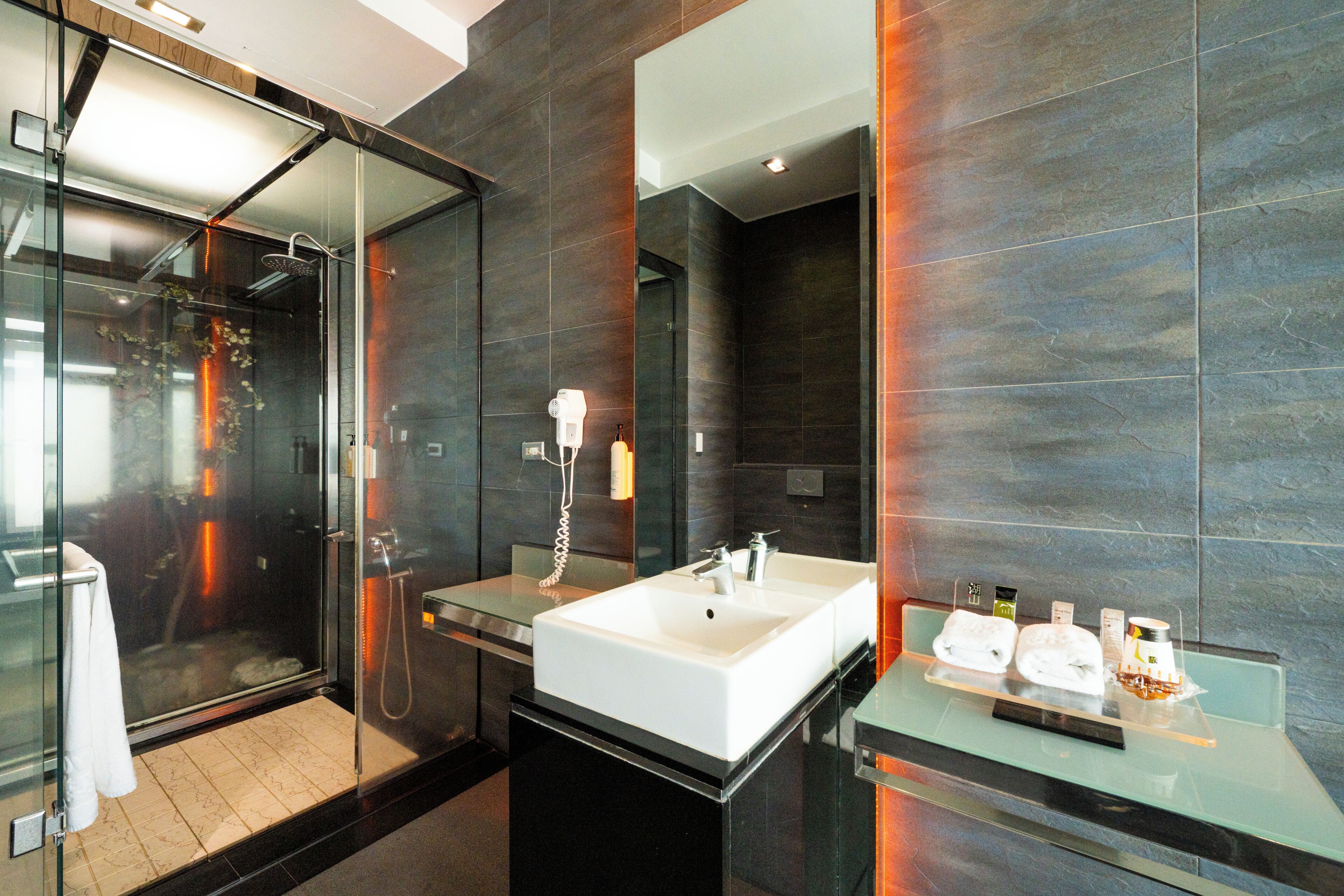 A modern bathroom with a glass shower enclosure, a large white sink, and a glass countertop with toiletries.