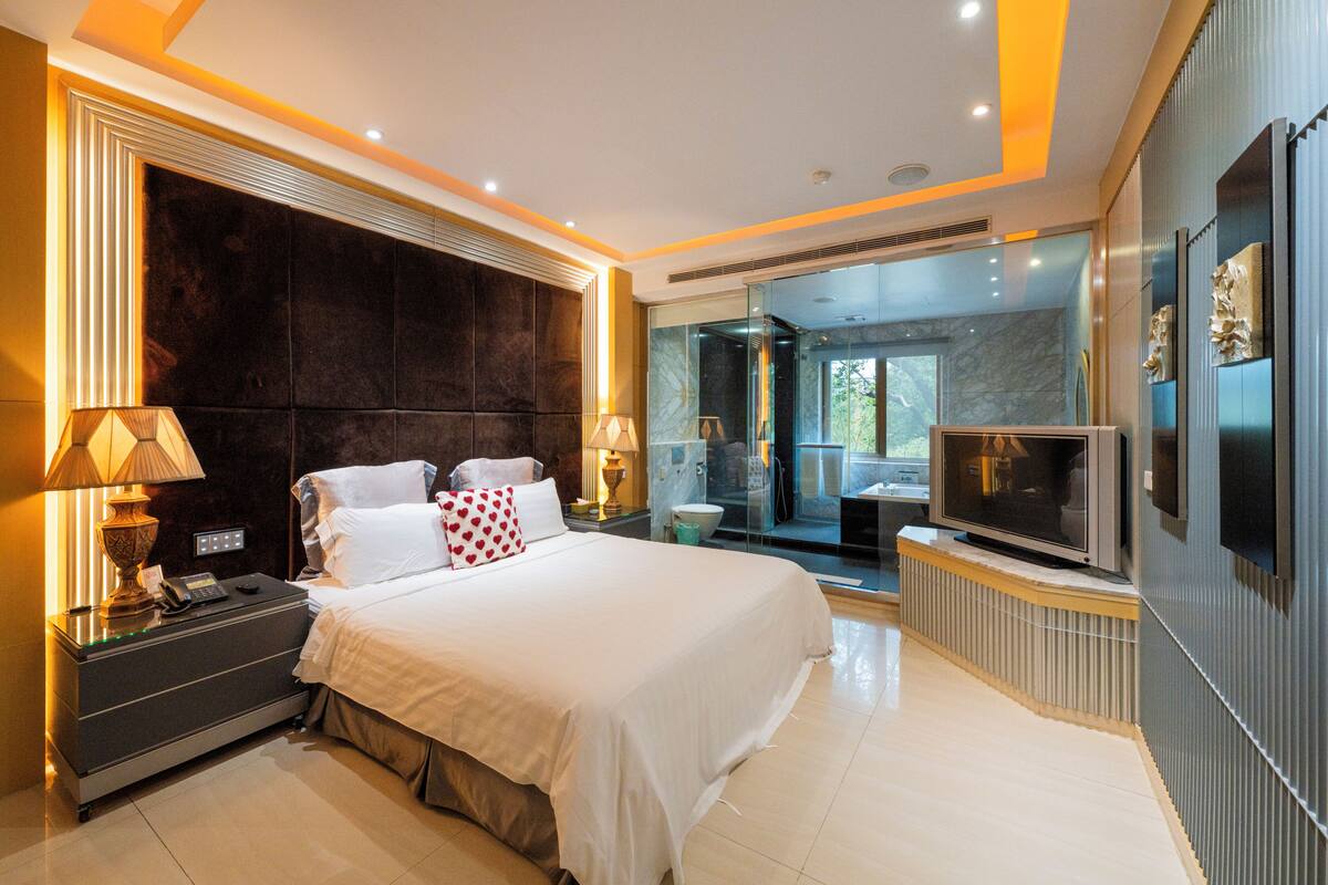 A modern hotel room with a large bed, bedside lamps, a TV, and a bathroom with a glass shower.