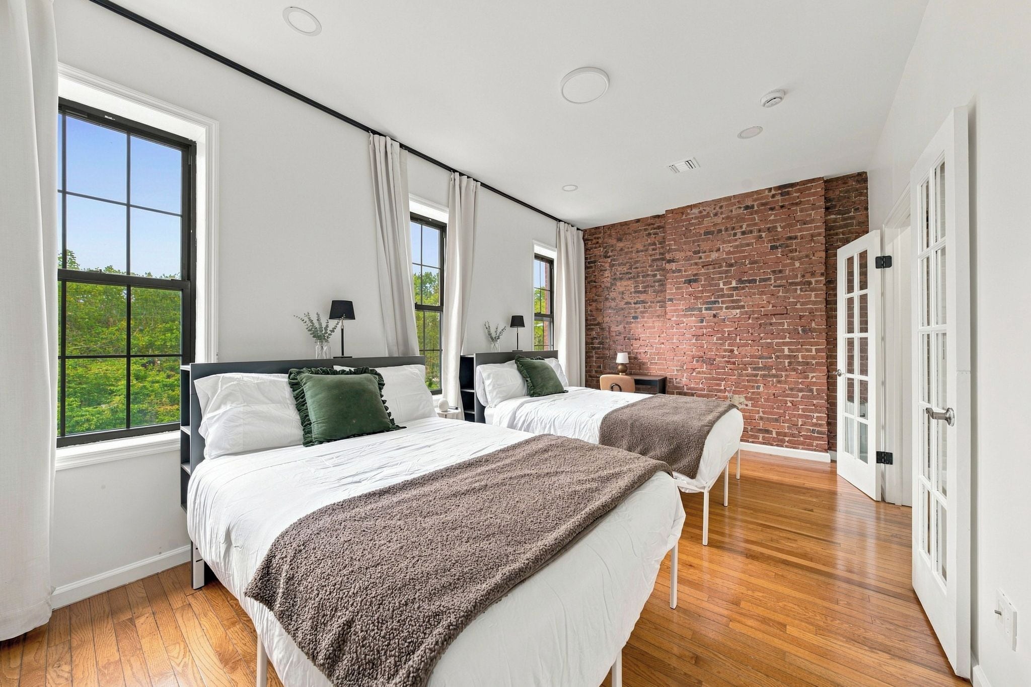 Quarto moderno com duas camas, janelas amplas, parede de tijolos e piso de madeira.