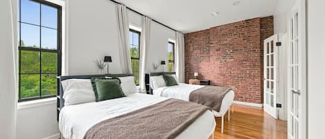 Quarto moderno com duas camas, janelas amplas, parede de tijolos e piso de madeira.