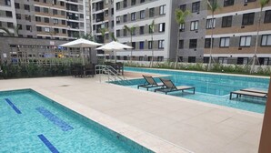 Een zwembad met ligbedden en parasols, omringd door een gebouw met meerdere balkons.