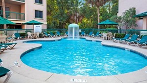 Piscina com espreguiçadeiras e guarda-sóis, cercada por uma vedação e árvores.